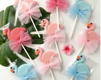 May include: A collection of flamingo-shaped lollipops in shades of pink and light blue, each individually wrapped in clear plastic and adorned with a matching tulle bow tied around the white stick, arranged on a large green monstera leaf.