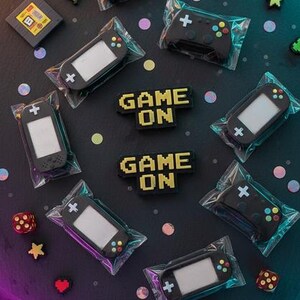May include: An assortment of gaming-themed items, including black game controllers and handheld consoles, packaged in clear plastic. The phrase "GAME ON" is displayed in pixelated yellow letters. Decorative dice, stars, and heart shapes are scattered around the items.