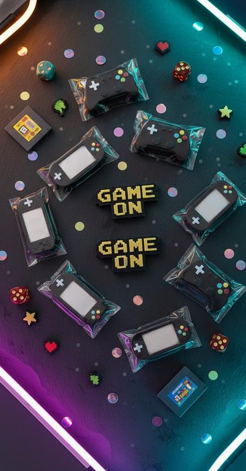 May include: An assortment of gaming-themed items, including black game controllers and handheld consoles, packaged in clear plastic. The phrase "GAME ON" is displayed in pixelated yellow letters. Decorative dice, stars, and heart shapes are scattered around the items.