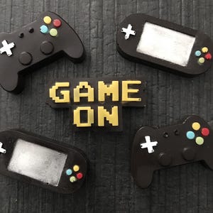 May include: A collection of black gaming-themed accessories, including two game controllers, two handheld game consoles, and a pixelated "GAME ON" sign in yellow. The controllers have colorful buttons and white cross-shaped directional pads.