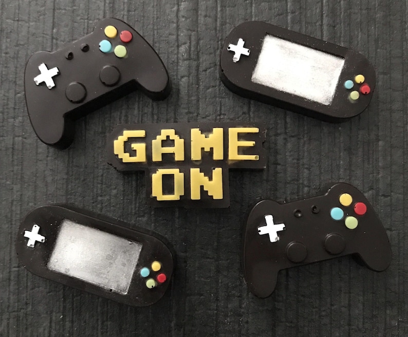 May include: A collection of black gaming-themed accessories, including two game controllers, two handheld game consoles, and a pixelated "GAME ON" sign in yellow. The controllers have colorful buttons and white cross-shaped directional pads.