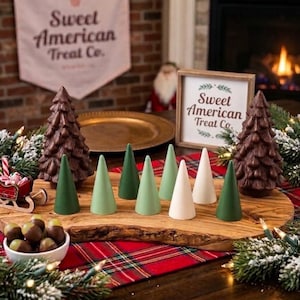 May include: A collection of decorative cone-shaped trees in shades of green and white, arranged on a wooden board alongside two textured chocolate-colored trees, a small bowl of chocolate candies, and a sign reading Sweet American Treat Co.