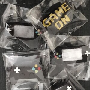 May include: Several black video game controller-shaped items, each individually packaged in clear plastic. The controllers have colorful buttons and a white cross-shaped directional pad. Some packages display the text "GAME ON" in pixelated gold letters.