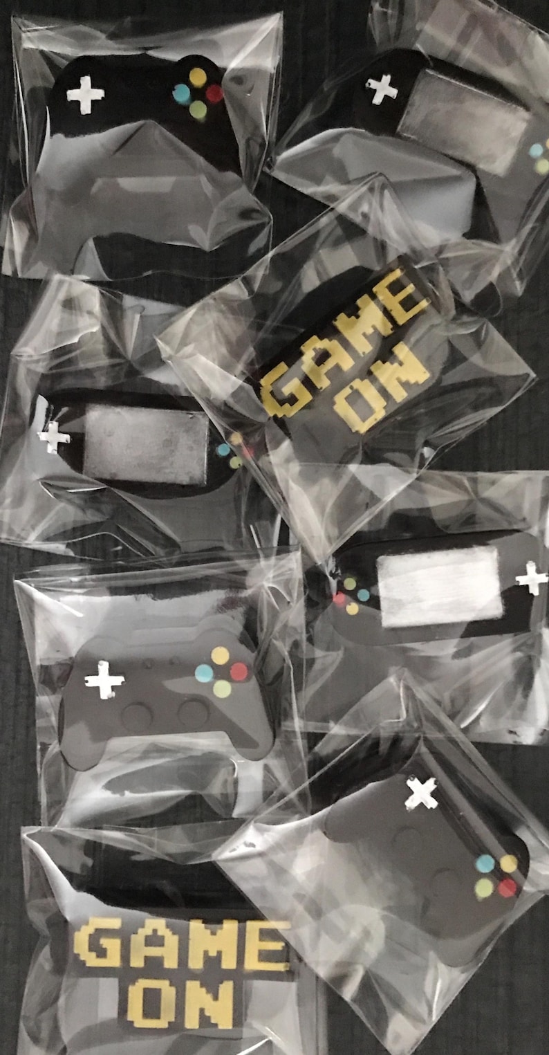 May include: Several black video game controller-shaped items, each individually packaged in clear plastic. The controllers have colorful buttons and a white cross-shaped directional pad. Some packages display the text "GAME ON" in pixelated gold letters.