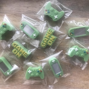 May include: A collection of individually wrapped, light green gaming-themed candies. The candies include game controllers, handheld game consoles, and blocks with the words "GAME ON" in yellow. The candies are on a light brown wooden surface.