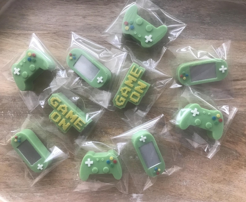 May include: A collection of individually wrapped, light green gaming-themed candies. The candies include game controllers, handheld game consoles, and blocks with the words "GAME ON" in yellow. The candies are on a light brown wooden surface.