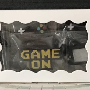 May include: A white box with a wavy cutout revealing a black gaming-themed design. The design includes a controller, cross buttons, and the words "GAME ON" in pixelated gold letters. The items are packaged in clear plastic.