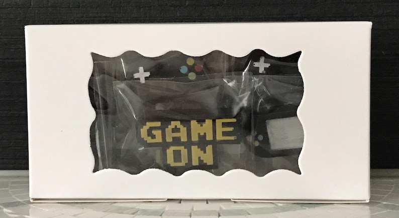 May include: A white box with a wavy cutout revealing a black gaming-themed design. The design includes a controller, cross buttons, and the words "GAME ON" in pixelated gold letters. The items are packaged in clear plastic.