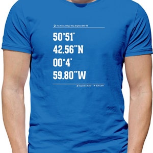 Könnte beinhalten: Ein königsblaues T-Shirt mit weißem Text, der "The Amex, Village Way, Brighton BN1 98" und Koordinaten lautet: "50°51′ 42.56"N 00°4′ 59.80"W". Der untere Teil des Shirts lautet "Capacity: 30,666 Built: 2011".