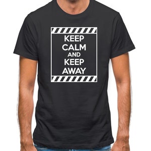 May include: Black t-shirt with the text "KEEP CALM AND KEEP AWAY" in a white rectangular frame. The text is centered and in a bold, sans-serif font. The frame has diagonal stripes at the top and bottom.