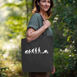 Könnte beinhalten: Schwarze Tragetasche mit weißer Grafik, die die menschliche Evolution darstellt und mit einer Curling-Figur endet. Die Tasche wird von einer Person in einem grünen Oberteil vor einem verschwommenen grünen Hintergrund gehalten.