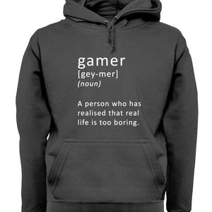May include: A dark gray hooded sweatshirt with a white graphic print that defines the word "gamer" as a person who has realized that real life is too boring.