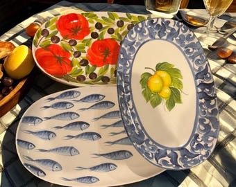 May include: Three decorative ceramic serving platters. One features red tomatoes and olives, another has blue fish, and the third has a lemon and leaf design with a blue border. Ideal for serving appetizers or displaying food.