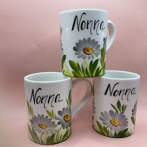 May include: Three white ceramic mugs with a floral design and the word "Nonna" written in black cursive. The mugs have a white background with grey and yellow flowers.