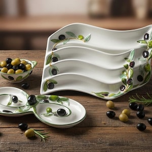 May include: White ceramic serving dishes with olive branch designs. The set includes a tiered server, a divided dish, and a small bowl. Black and green olives are scattered on a wooden surface.