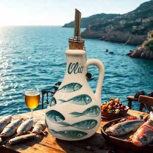 Puede incluir: Una aceitera de cer&aacute;mica blanca con asa, decorada con ilustraciones de peces azules y la palabra Olio en letra cursiva. La botella cuenta con un tap&oacute;n de corcho rematado con un vertedor de acero inoxidable para servir aceite o l&iacute;quidos.