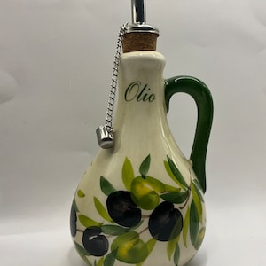 Puede incluir: Una aceitera de cer&aacute;mica color crema con un dise&ntilde;o pintado a mano de aceitunas negras y verdes con hojas. La botella tiene un asa verde, la palabra "Olio" impresa en letra cursiva negra y un vertedor de acero inoxidable con corcho y cadena.