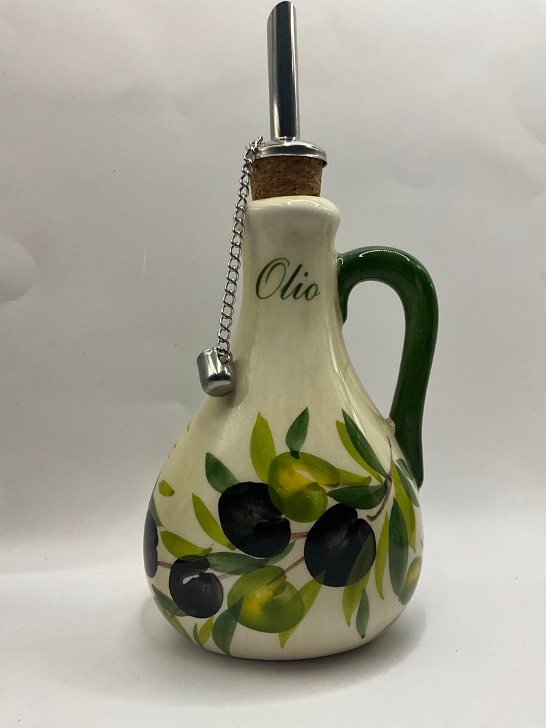 Puede incluir: Una aceitera de cer&aacute;mica color crema con un dise&ntilde;o pintado a mano de aceitunas negras y verdes con hojas. La botella tiene un asa verde, la palabra "Olio" impresa en letra cursiva negra y un vertedor de acero inoxidable con corcho y cadena.