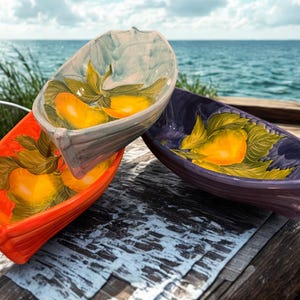 May include: Three ceramic boat-shaped bowls in orange, gray, and purple. Each bowl features a hand-painted design of yellow pears with green leaves. The bowls are displayed on a wooden surface with a blurred ocean background.