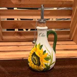 Puede incluir: Una aceitera de cer&aacute;mica blanca con un dise&ntilde;o de girasol amarillo pintado a mano y hojas verdes. La botella tiene un asa verde, un tap&oacute;n de corcho con un vertedor de acero inoxidable y una peque&ntilde;a cadena met&aacute;lica. La palabra Olio est&aacute; impresa.