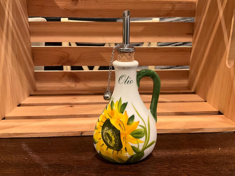 Puede incluir: Una aceitera de cer&aacute;mica blanca con un dise&ntilde;o de girasol amarillo pintado a mano y hojas verdes. La botella tiene un asa verde, un tap&oacute;n de corcho con un vertedor de acero inoxidable y una peque&ntilde;a cadena met&aacute;lica. La palabra Olio est&aacute; impresa.