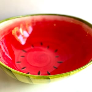 May include: A red and green ceramic bowl with a watermelon design. The bowl has a green rim and black seeds painted on the inside.