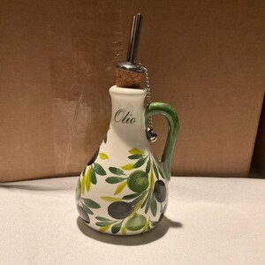 Puede incluir: Una aceitera de cer&aacute;mica blanca con asa verde y un patr&oacute;n pintado a mano de ramas de olivo con aceitunas negras y verdes. La palabra Olio est&aacute; impresa en verde en el cuello. Incluye un tap&oacute;n de corcho con un vertedor de metal y una cadena.