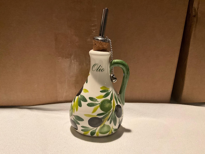 Puede incluir: Una aceitera de cer&aacute;mica blanca con asa verde y un patr&oacute;n pintado a mano de ramas de olivo con aceitunas negras y verdes. La palabra Olio est&aacute; impresa en verde en el cuello. Incluye un tap&oacute;n de corcho con un vertedor de metal y una cadena.