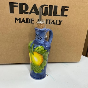 May include: A blue ceramic oil cruet featuring a hand-painted yellow lemon and green leaves design. The bottle has a cork stopper with a silver-colored metal pour spout and a small handle. The word Olio is written in black script on the neck.