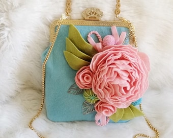May include: A light blue handbag with a gold-colored frame and handle. The bag is decorated with a large pink felt flower, green leaves, and small pink accents. A gold chain strap is attached.