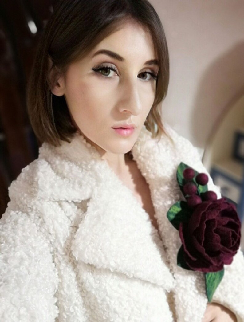 Puede incluir: Una chaqueta de piel sint&eacute;tica blanca con un broche de flor de fieltro burdeos. La flor tiene un tallo y hojas verdes.