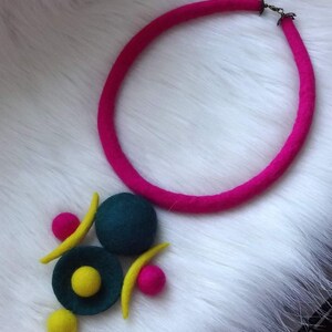 May include: A necklace featuring a thick, bright pink felted cord with a metal clasp. The pendant consists of a large dark teal felt sphere, a smaller dark teal felt disc with a yellow center, two yellow felt crescents, and two small pink felt spheres.