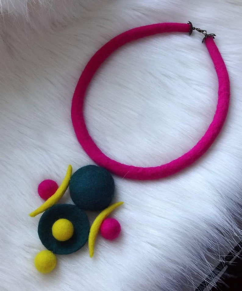May include: A necklace featuring a thick, bright pink felted cord with a metal clasp. The pendant consists of a large dark teal felt sphere, a smaller dark teal felt disc with a yellow center, two yellow felt crescents, and two small pink felt spheres.