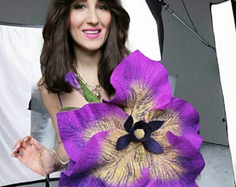 May include: A woman wearing a purple dress with a large purple and yellow felt flower attached to the front. The flower has a black center and a gold chain is draped around her neck.