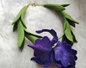 Puede incluir: Una flor de fieltro morado con hojas de fieltro verde, elaborada en forma de collar. La flor tiene un diseño delicado y en capas, y las hojas están dispuestas en cascada.