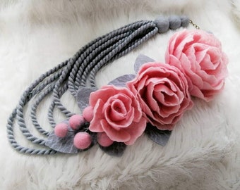 Könnte beinhalten: Eine graue Filzkette mit mehreren Strängen und einer rosa Filzblumenverzierung. Die Blume hat drei Lagen Blütenblätter und ist mit grauen Filzblättern verziert.