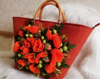 以下が含まれることがあります： オレンジ色のフェルトの花のアップリケが付いた赤いフェルトのトートバッグ。花はたくさんの花びらでできており、緑色のフェルトの葉とベリーが付いています。
