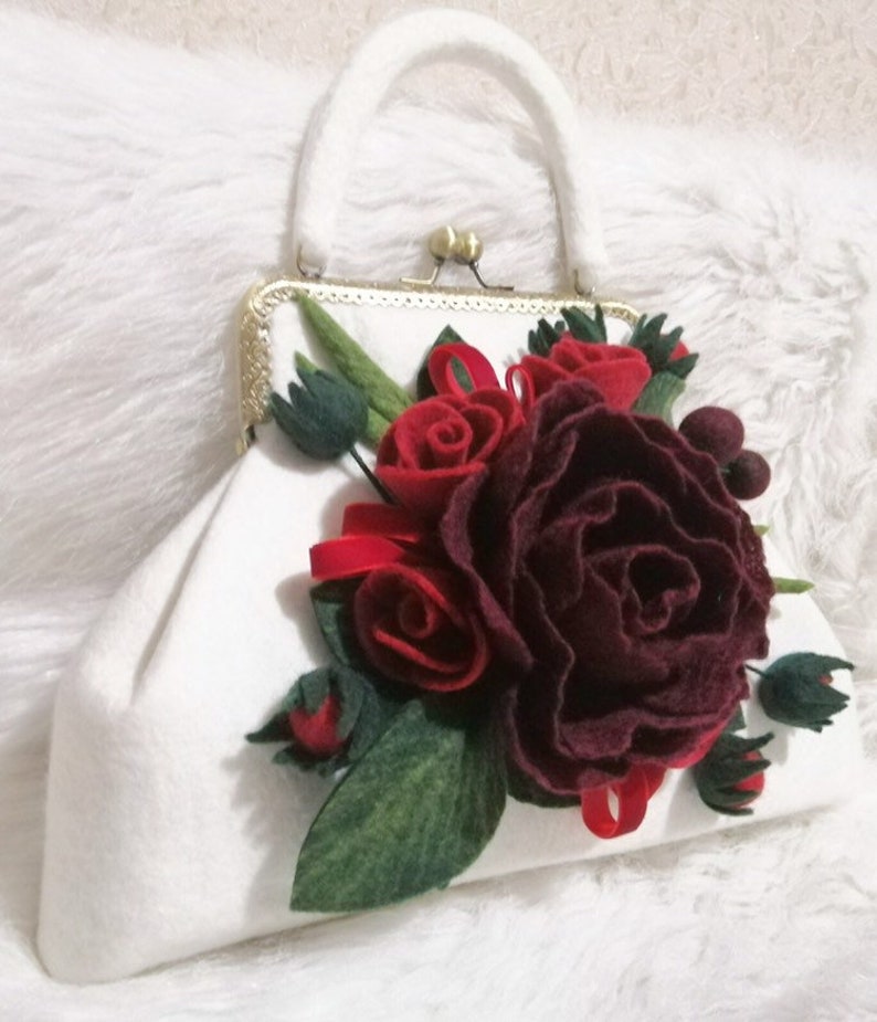 Puede incluir: Un bolso de mano de fieltro blanco con un cierre dorado y una gran flor de fieltro burdeos y roja.