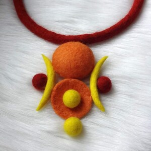 May include: A felted wool necklace featuring a thick red cord and a central pendant composed of a large orange sphere, a flat orange disc with a small yellow sphere in the center, two yellow crescent shapes, and two small red spheres.