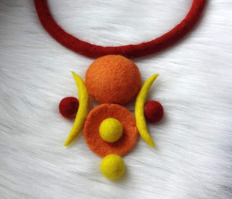 May include: A felted wool necklace featuring a thick red cord and a central pendant composed of a large orange sphere, a flat orange disc with a small yellow sphere in the center, two yellow crescent shapes, and two small red spheres.