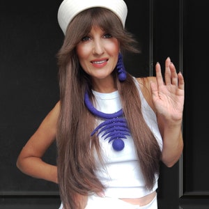 May include: A woman wearing a white sailor hat, a white sleeveless top, and white pants. She is wearing a large, blue felt necklace and matching earrings. She is smiling and waving.