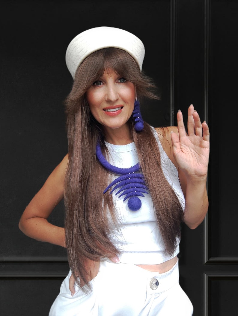 May include: A woman wearing a white sailor hat, a white sleeveless top, and white pants. She is wearing a large, blue felt necklace and matching earrings. She is smiling and waving.