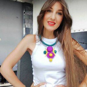 May include: A white sleeveless top paired with a felted necklace featuring a teal cord and a central pendant composed of purple and yellow circular and crescent-shaped elements. The necklace includes small teal and yellow spheres as decorative accents.