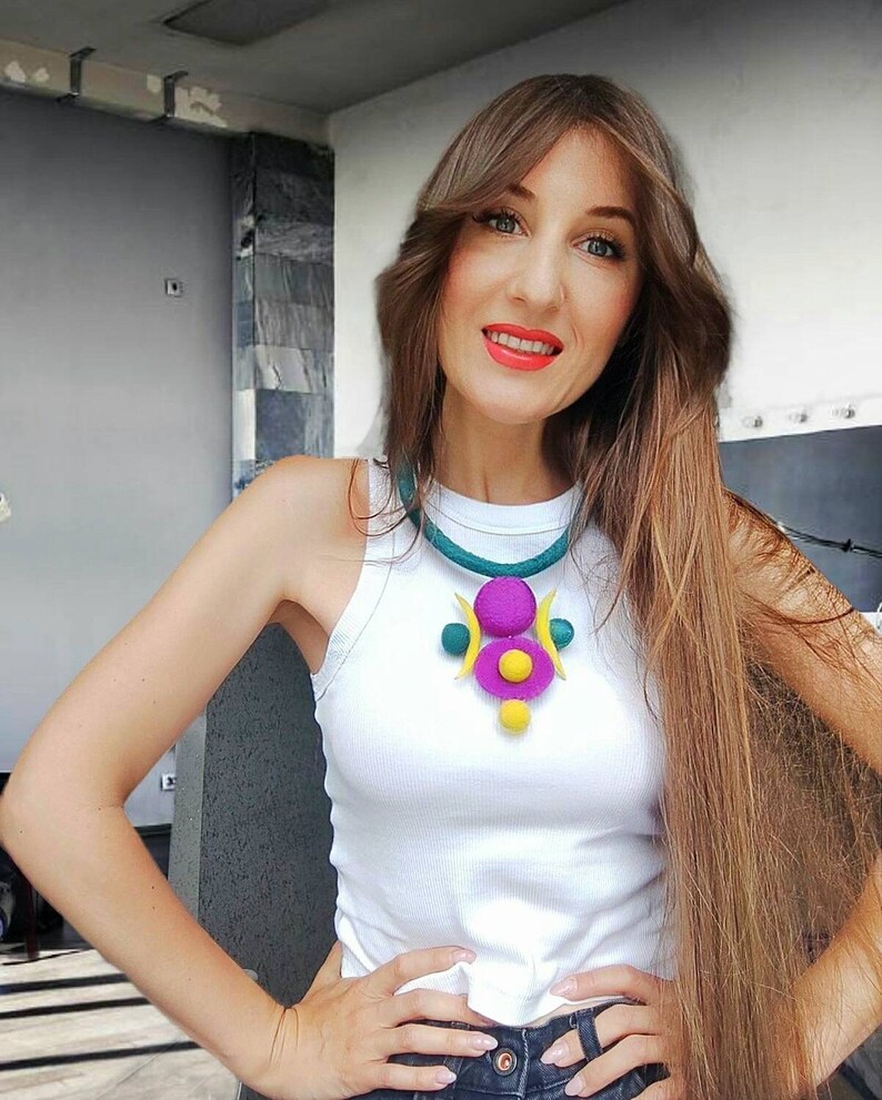 May include: A white sleeveless top paired with a felted necklace featuring a teal cord and a central pendant composed of purple and yellow circular and crescent-shaped elements. The necklace includes small teal and yellow spheres as decorative accents.