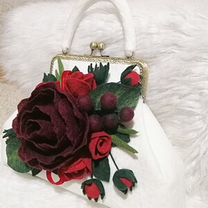 Puede incluir: Un bolso de mano de fieltro blanco con cierre dorado y una decoraci&oacute;n de flores de fieltro burdeos y rojo.