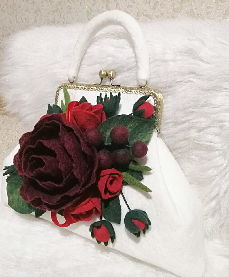 Puede incluir: Un bolso de mano de fieltro blanco con cierre dorado y una decoraci&oacute;n de flores de fieltro burdeos y rojo.