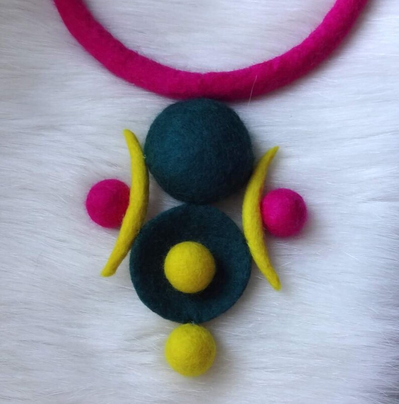 May include: A necklace featuring a thick, bright pink felt cord and a central pendant composed of a large dark teal felt sphere, a smaller dark teal felt disc with a yellow felt sphere in the center, two yellow felt crescents, and two pink spheres.
