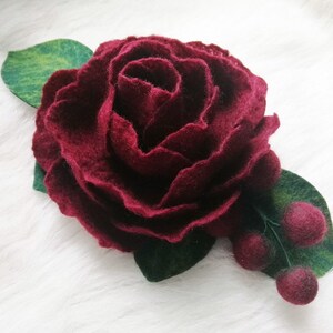 Puede incluir: Una flor de fieltro burdeos con hojas de fieltro verde y bayas de fieltro burdeos.