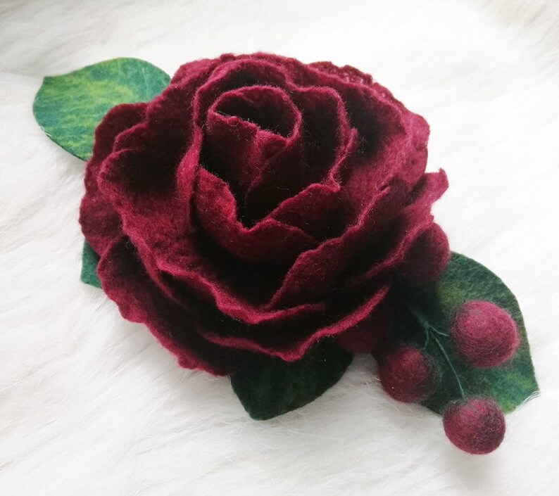 Puede incluir: Una flor de fieltro burdeos con hojas de fieltro verde y bayas de fieltro burdeos.