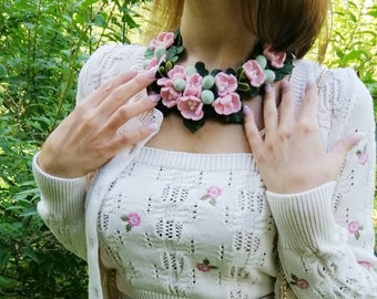 Könnte beinhalten: Eine florale Halskette mit rosa Blüten, grünen Blättern und kleinen grünen Kugeln. Die Halskette wird mit einem cremefarbenen Strickoberteil und einer Strickjacke getragen. Der Hintergrund ist ein grüner Garten.